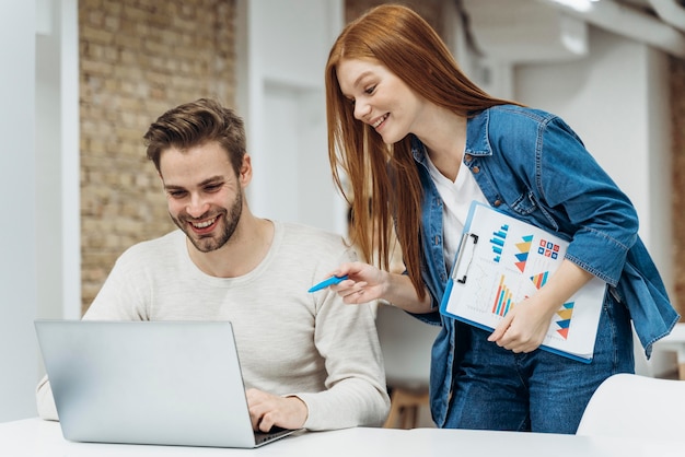 Two coworkers smiling while collaborating on a laptop, with one holding a clipboard showing colorful charts and graphs.